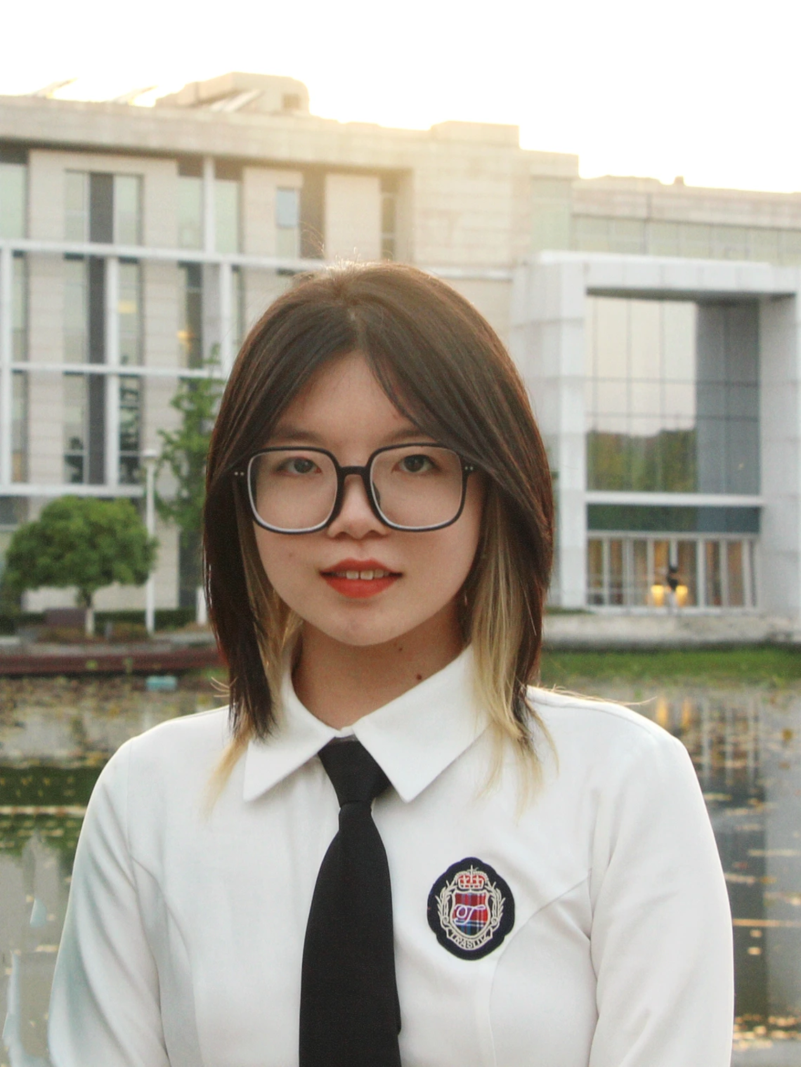 Jialij Li stands wearing a shirt and tie in front of a lake and building with lots of glass doors and windows in the background