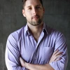 Professional photo of Lucas Belury standing in front of a matte background wearing a collared buttondown shirt with his arms crossed