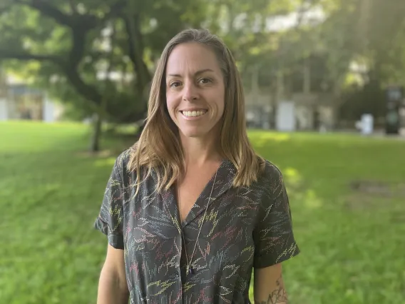 Dr. Megan Carney stands smiling in front of a background of grass, trees, and sunlight.