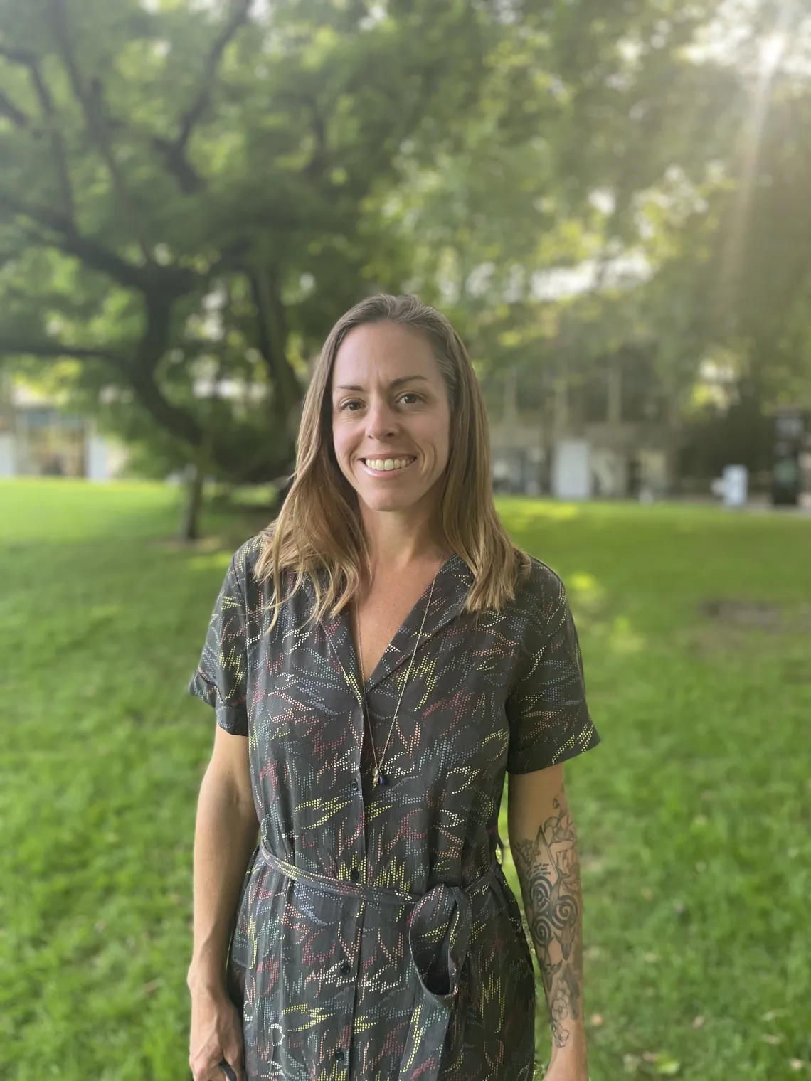 Dr. Megan Carney stands smiling in front of a background of grass, trees, and sunlight.